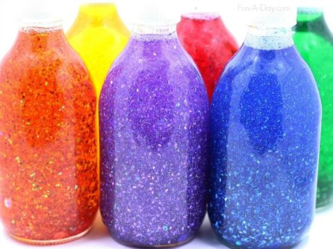 Five glass bottles filled with glittery liquid are lined up. The contents are vibrant orange, yellow, purple, blue, and green. The bottles are close together, showcasing a colorful spectrum. Labels or backgrounds are not visible.