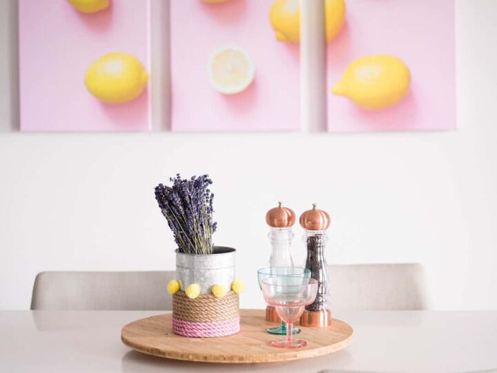 A dining table with a wooden tray holding lavender in a tin can, two glasses, and copper salt and pepper grinders. Three wall art pieces above feature yellow lemons on a pink background. White chairs surround the table.