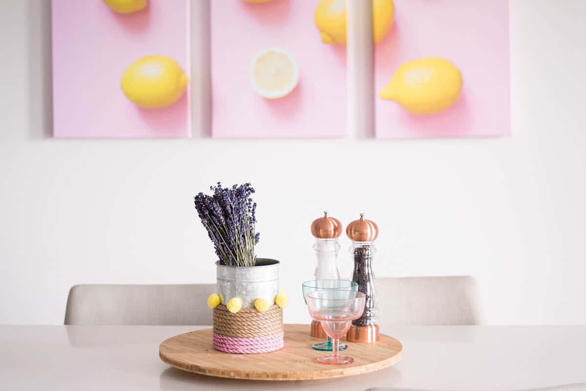 A dining table with a wooden tray holding lavender in a tin can, two glasses, and copper salt and pepper grinders. Three wall art pieces above feature yellow lemons on a pink background. White chairs surround the table.
