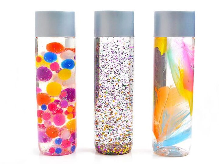 Three clear sensory bottles lined up, each with a blue cap. The first contains colorful pom-poms, the second is filled with glitter, and the third has vibrant feathers inside.