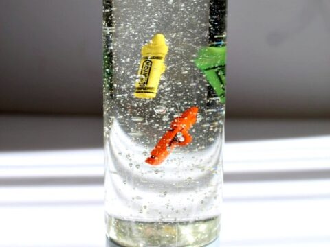 A clear bottle with a gray base, filled with liquid and glitter. Inside, an orange fish-shaped object and a yellow crayon float. The bottle is illuminated by sunlight, casting shadows on the striped surface below.