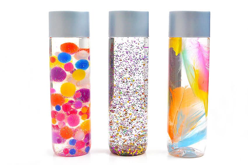 Three clear sensory bottles lined up, each with a blue cap. The first contains colorful pom-poms, the second is filled with glitter, and the third has vibrant feathers inside.