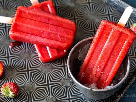 Three vibrant red strawberry popsicles on a textured dark plate, with whole strawberries scattered around. One popsicle is placed in a small black cup filled with ice, glistening with moisture.