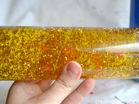 A hand holding a clear bottle filled with yellow liquid and suspended gold glitter. The background is blurred, focusing attention on the bottle’s contents.