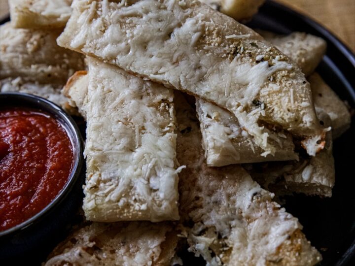 A plate of cheesy breadsticks is stacked on a dark plate, accompanied by a small black bowl of red marinara sauce. The breadsticks are topped with melted cheese and have a golden-brown crust. The background is a wooden surface.