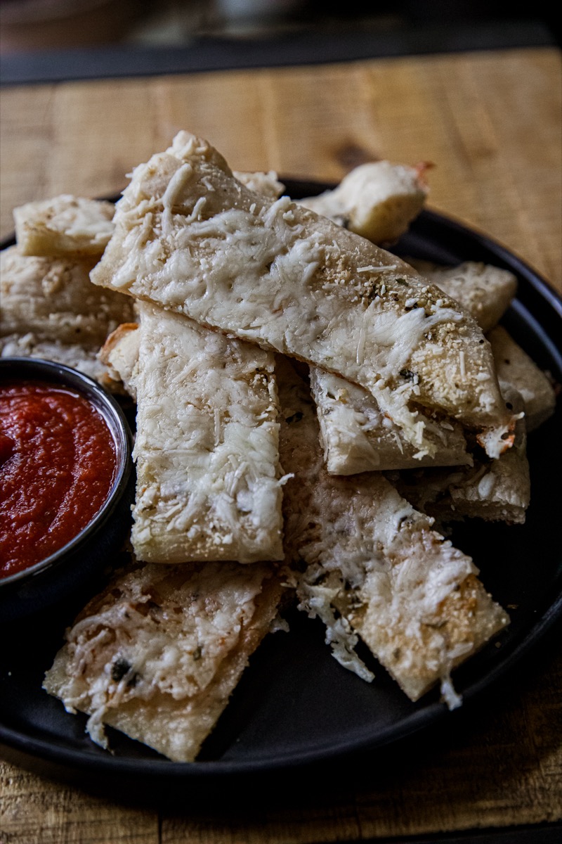 A plate of cheesy breadsticks is stacked on a dark plate, accompanied by a small black bowl of red marinara sauce. The breadsticks are topped with melted cheese and have a golden-brown crust. The background is a wooden surface.