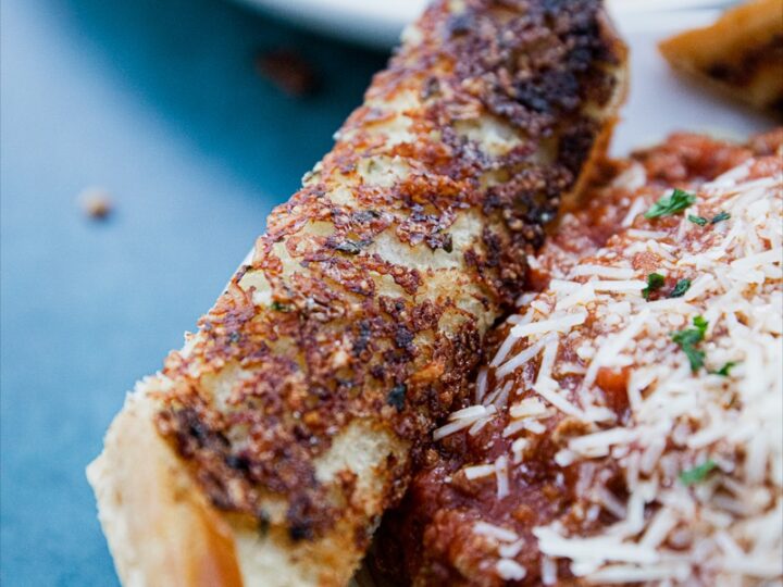 Close-up of a plate with spaghetti covered in meat sauce and grated cheese, accompanied by a piece of crispy garlic bread. The dish is served on a white plate against a blue surface.