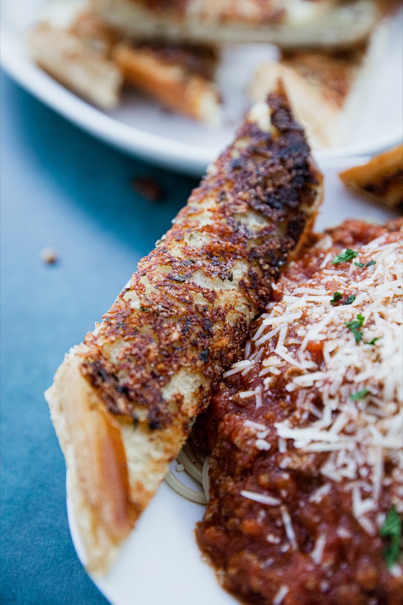 Close-up of a plate with spaghetti covered in meat sauce and grated cheese, accompanied by a piece of crispy garlic bread. The dish is served on a white plate against a blue surface.
