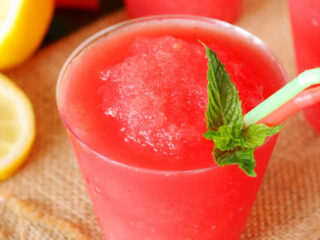 A glass of bright red slushie with a green and red straw, garnished with a mint sprig. Its sitting on a burlap cloth, next to a lemon slice, with more slushies visible in the background.