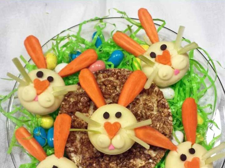 Five cheese bites decorated as bunny faces with almond ears and carrot noses, placed around a central stack of cookies. They rest on a bed of green shredded paper with colorful jelly beans scattered around. Text reads Babybel Bunnies.
