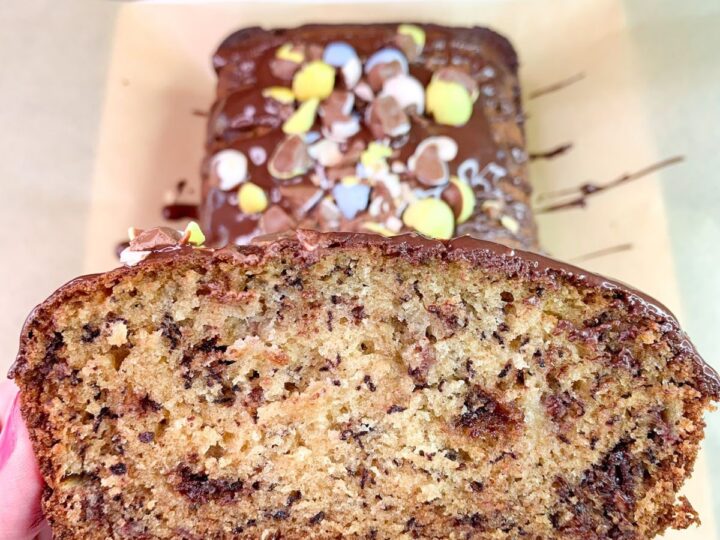 A hand holds a slice of banana bread with chocolate chips, revealing its moist, speckled interior. The loaf is topped with chocolate drizzle and colorful candy pieces, resting on parchment paper with a blue-striped cloth nearby.