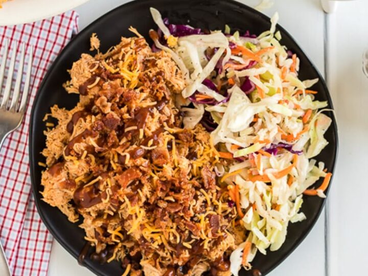 A black plate filled with shredded chicken topped with cheese and crispy bits, drizzled with sauce, next to a side of coleslaw. The plate is set on a white table with a fork, napkin, and a sauce dish nearby.
