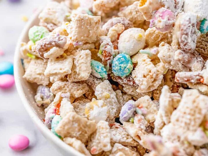 A close-up of a bowl filled with a colorful snack mix. It includes cereals, coated in powdered sugar, with pastel-colored candies and pretzel pieces. Some yellow and pink sprinkles are scattered nearby on the surface.