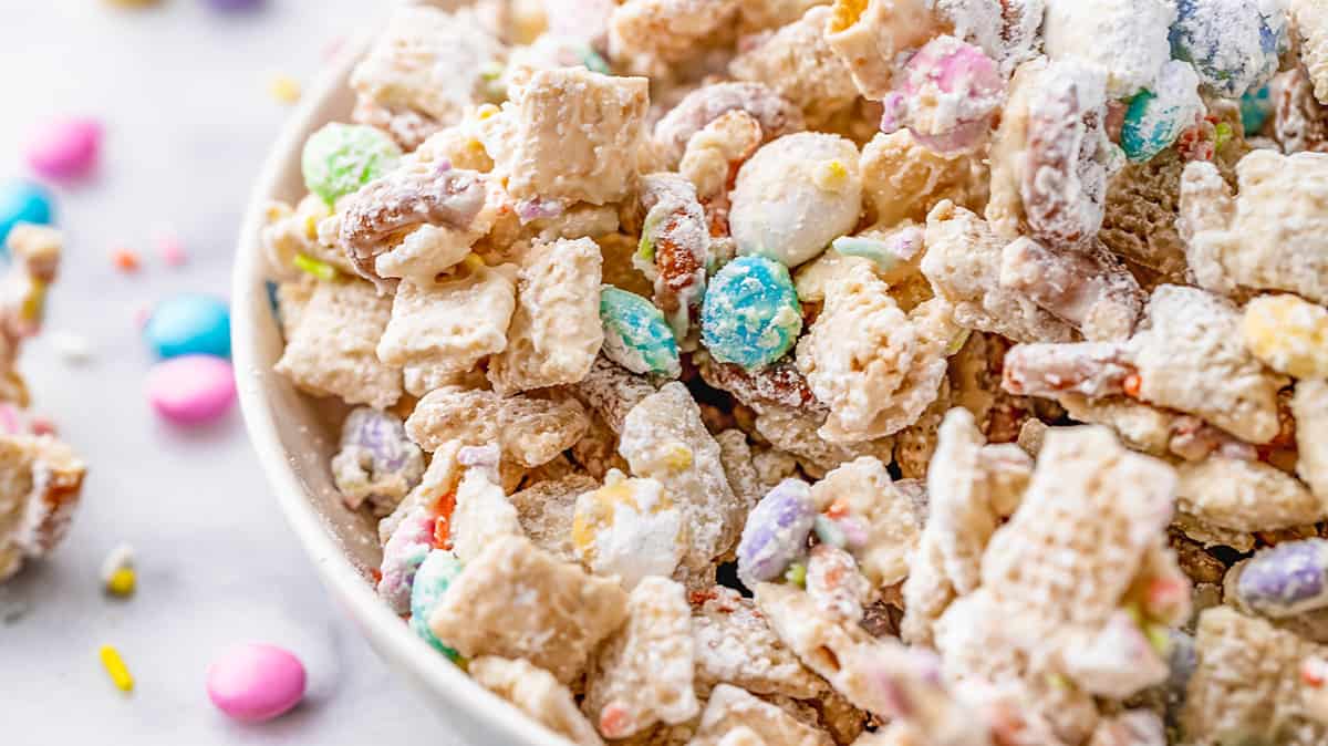 A close-up of a bowl filled with a colorful snack mix. It includes cereals, coated in powdered sugar, with pastel-colored candies and pretzel pieces. Some yellow and pink sprinkles are scattered nearby on the surface.
