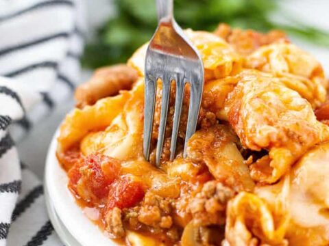 A fork is being used to pick up tortellini from a plate. The pasta is mixed with a rich tomato and meat sauce. A striped cloth napkin is visible, and there are blurred greens in the background.