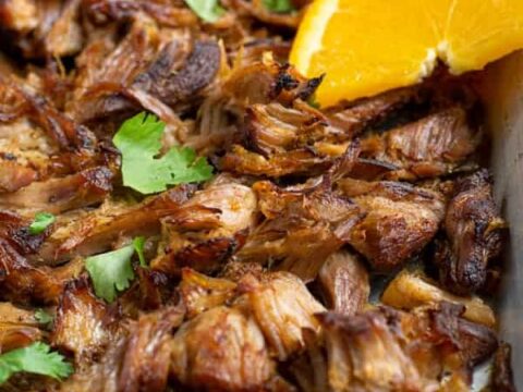 Close-up of crispy, shredded carnitas garnished with cilantro leaves. An orange slice and lime wedge are placed on top for garnish. The meat is golden brown with slightly charred edges, suggesting its freshly cooked and flavorful.