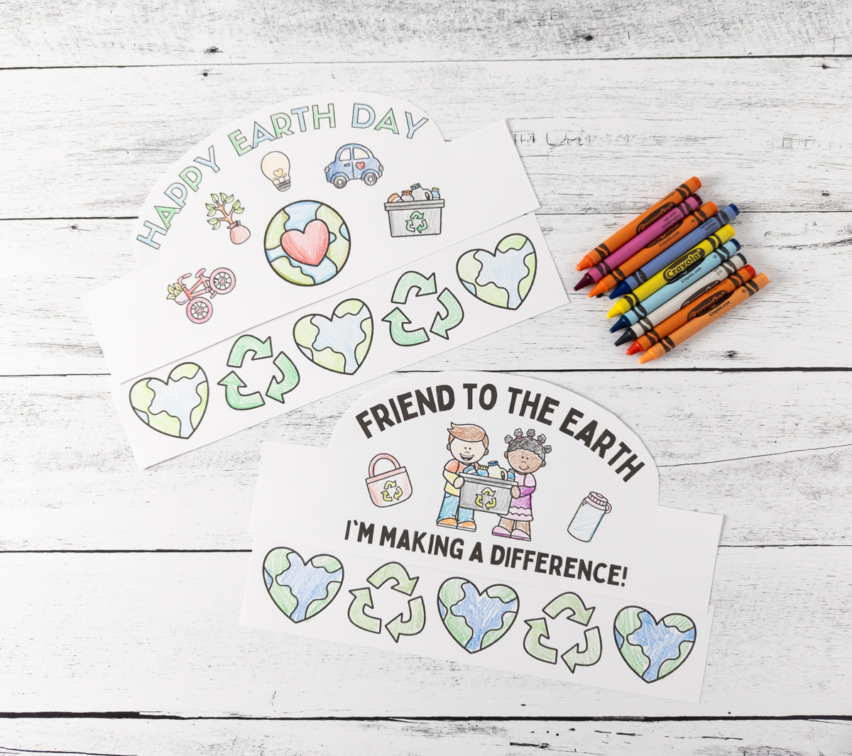 Two Earth Day-themed coloring sheets with drawings of hearts, recycling symbols, and Earths. The top sheet says Happy Earth Day, and the bottom says Friend to the Earth Im Making a Difference! Crayons are beside the sheets on a wooden surface.