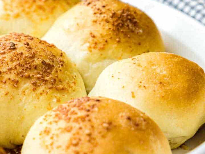 Freshly baked dinner rolls in a white baking dish, topped with a sprinkle of seasoning. A stack of white plates and a checkered cloth are visible in the background.