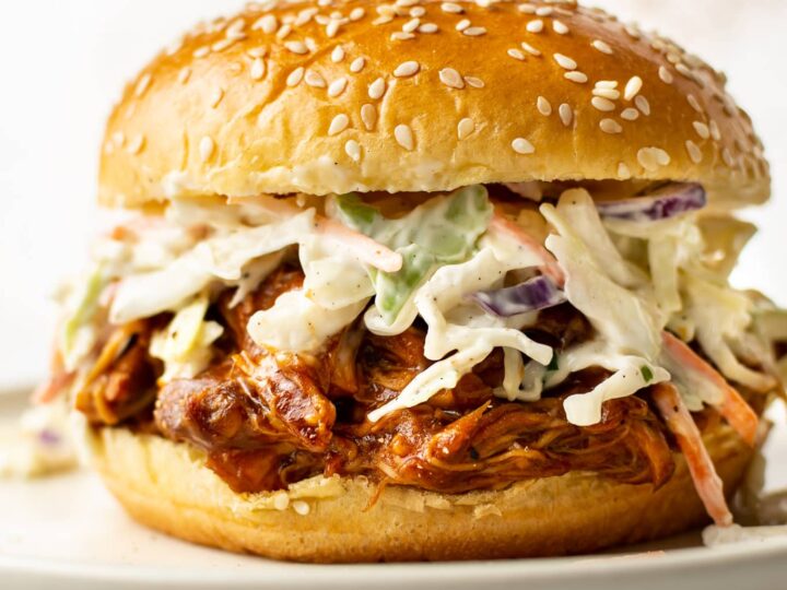 A pulled pork sandwich with coleslaw, served on a sesame seed bun, sits on a plate. The fillings overflow slightly, showcasing the vibrant colors of the coleslaw and the saucy pork.