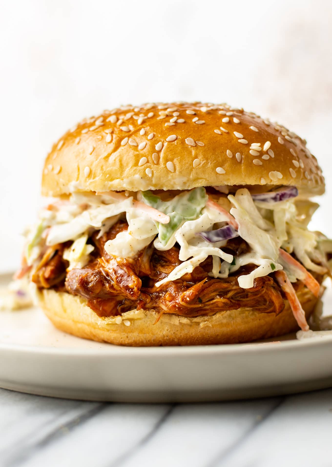 A pulled pork sandwich with coleslaw, served on a sesame seed bun, sits on a plate. The fillings overflow slightly, showcasing the vibrant colors of the coleslaw and the saucy pork.