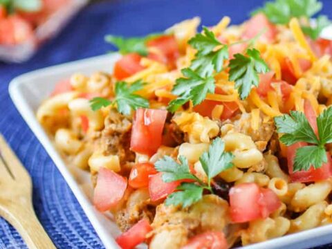 A bowl of macaroni pasta topped with diced tomatoes, cheddar cheese, and parsley on a blue woven placemat. A wooden fork is placed beside the bowl. A dish with more tomatoes is visible in the background.