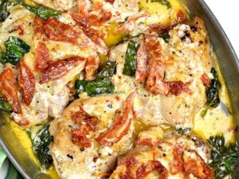 A dish of creamy Tuscan chicken in a pan, featuring chicken breasts topped with a rich sauce, sun-dried tomatoes, and wilted spinach. The golden-brown chicken is garnished with black pepper and herbs, surrounded by a creamy yellow sauce.