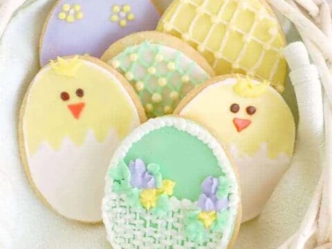 A basket containing decorated Easter cookies. The cookies are shaped like eggs and feature designs including chicks, flowers, and pastel patterns in yellow, green, purple, and white icing.