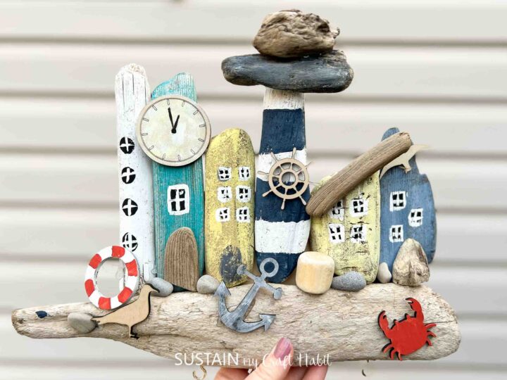 Driftwood artwork featuring painted wooden pieces as coastal buildings, a lighthouse, and nautical elements like a clock, anchor, lifebuoy, ship wheel, seagull, and crab. Accented with stones and a large wooden piece on top.