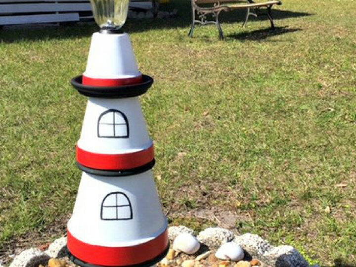 A decorative lighthouse made from stacked terra cotta pots is painted in red, white, and black. It sits on a bed of rocks with seashells around it. A lamp is on top. A park bench and trees are in the background. Text reads, DIY Terra Cotta Lighthouse.