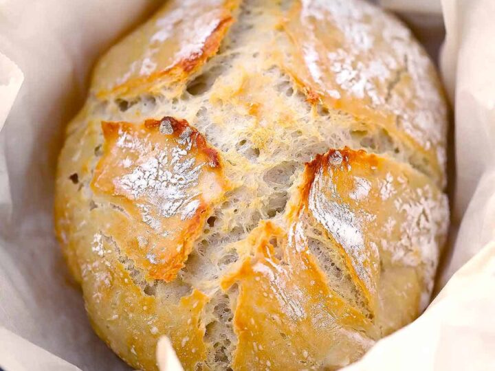 A freshly baked round loaf of bread with a golden, crusty top sits on parchment paper. The bread has a rustic look with a few cracks on the surface, revealing a soft, airy interior.