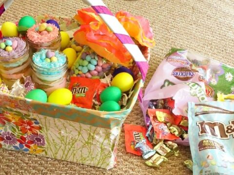 A colorful Easter basket filled with jars of layered desserts topped with mini eggs, surrounded by plastic eggs and candy wrappers. Next to it are bags of assorted candies like Snickers and M&Ms scattered on a carpeted floor.