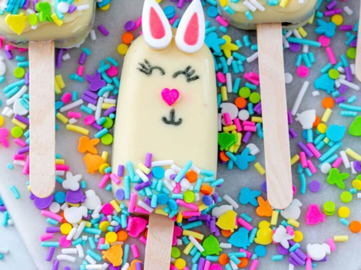 A white popsicle with a bunny face design lies on a tray, surrounded by colorful sprinkles and other popsicles. The bunny has pink ears, a pink heart-shaped nose, and closed eyes with eyelashes, with sprinkles on the lower half.