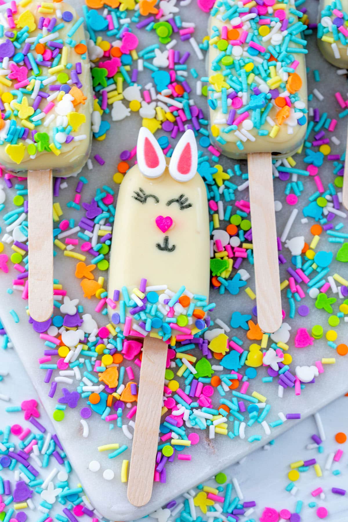 A white popsicle with a bunny face design lies on a tray, surrounded by colorful sprinkles and other popsicles. The bunny has pink ears, a pink heart-shaped nose, and closed eyes with eyelashes, with sprinkles on the lower half.