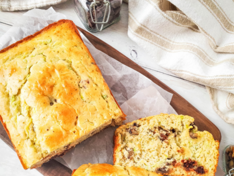 A loaf of zucchini bread with two slices cut, displayed on a wooden board with parchment paper. The text at the top reads Bisquick Zucchini Bread. A striped cloth and a jar are in the background. Text at the bottom: www.mommalew.com.