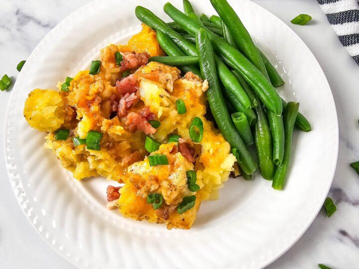 A white plate with a serving of cheesy mashed potatoes topped with bacon and chopped green onions, next to a portion of steamed green beans. The plate is on a marble surface near a striped cloth and scattered green onions.