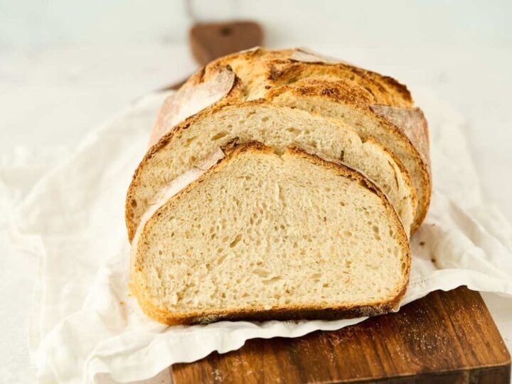 Sliced loaf of crusty bread on a wooden board with a white cloth underneath, displayed against a light background.