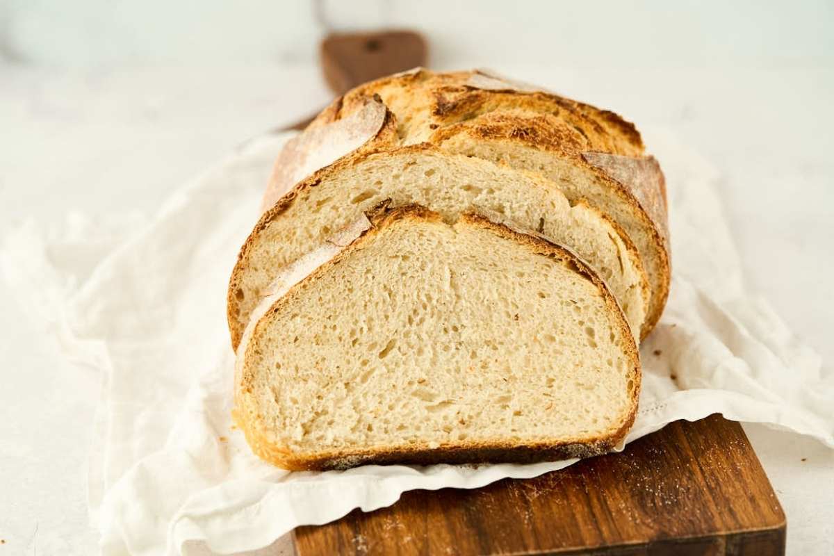 Sliced loaf of crusty bread on a wooden board with a white cloth underneath, displayed against a light background.