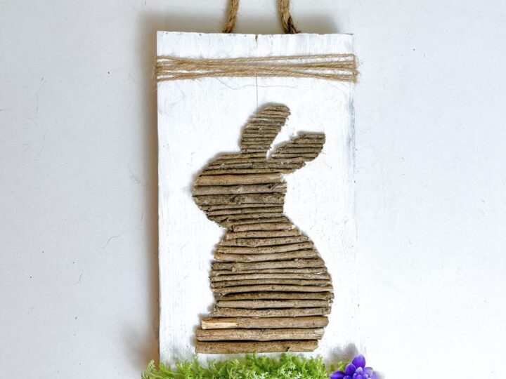 A rustic wall hanging features the silhouette of a rabbit made from vertical twigs on a white wooden background. It is adorned with twine at the top and faux green foliage with purple flowers at the bottom.