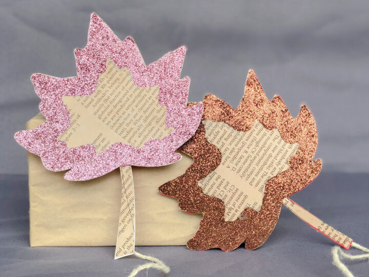 Two decorative leaves made from book pages and covered with pink and copper glitter are attached to sticks. They lean against a brown paper-wrapped box on a gray background.