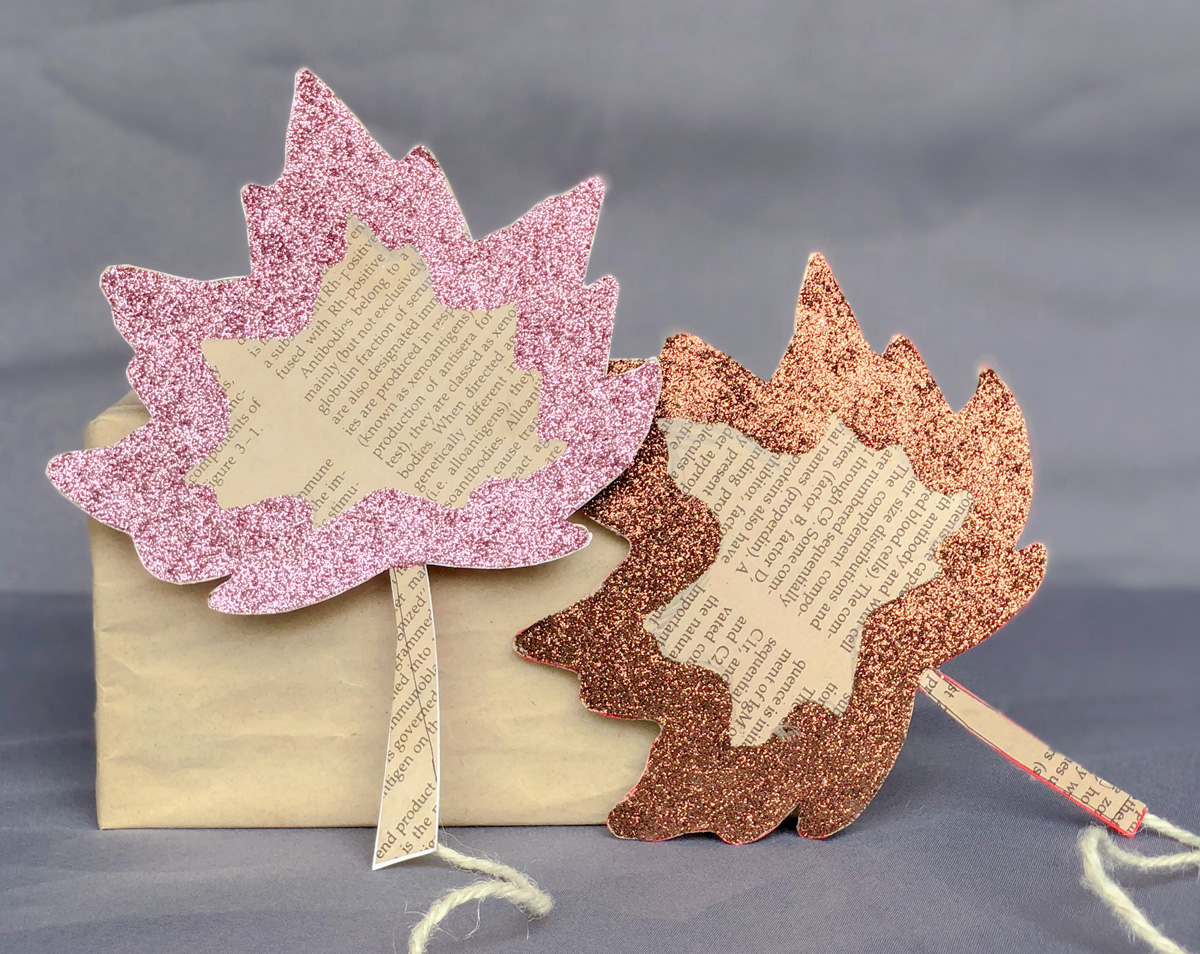 Two decorative leaves made from book pages and covered with pink and copper glitter are attached to sticks. They lean against a brown paper-wrapped box on a gray background.