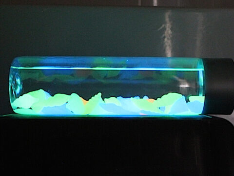A capsule-shaped glass tube containing glowing neon-colored materials in shades of blue, green, and yellow rests on a dark surface. This striking sensory bottle idea showcases its contents illuminated brightly against a dim background.