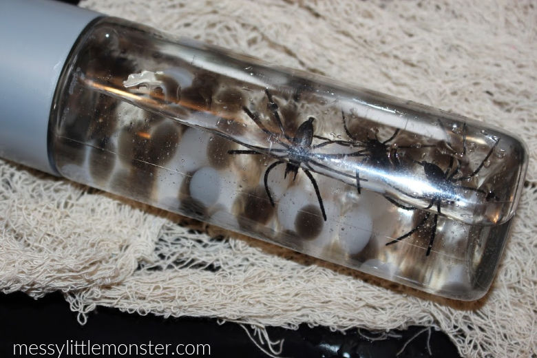 A clear bottle filled with water contains small toy spiders and round objects resembling eggs. The bottle is placed on a textured white cloth. The website name messylittlemonster.com is visible in the lower corner.