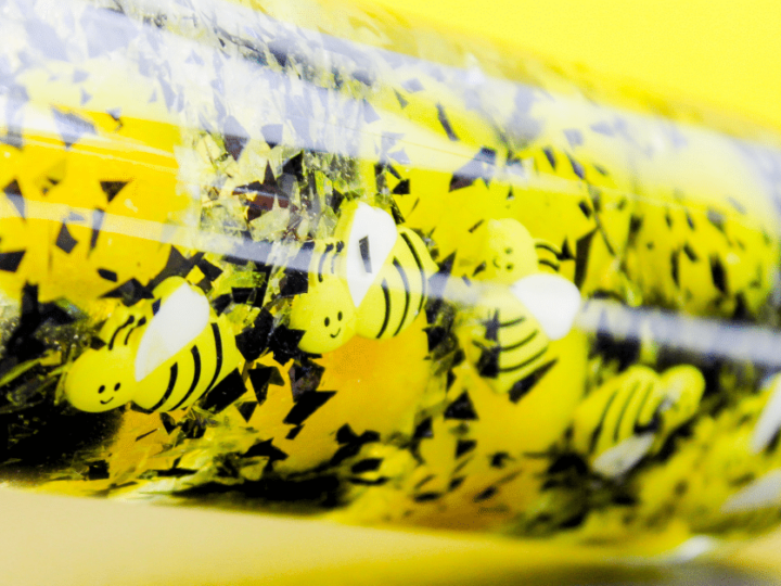 A close-up of a clear sensory bottle filled with yellow liquid, black and silver glitter, and small cartoon bee cutouts. The bright yellow background enhances the playful theme. The bottle is labeled Fun A Day in the bottom left corner.