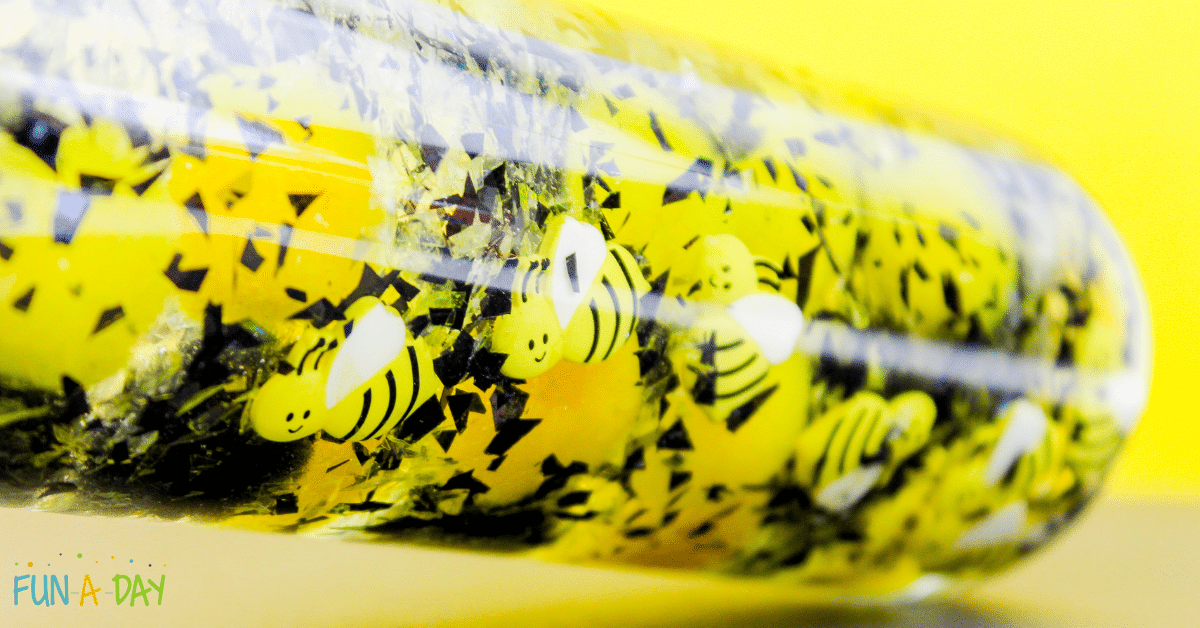 A close-up of a clear sensory bottle filled with yellow liquid, black and silver glitter, and small cartoon bee cutouts. The bright yellow background enhances the playful theme. The bottle is labeled Fun A Day in the bottom left corner.
