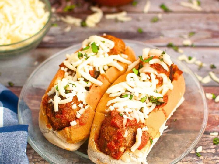 Two meatball subs on a glass plate, topped with melted cheese and garnished with chopped herbs. The wooden table is scattered with extra cheese and herbs. A bowl of shredded cheese, a jar of sauce, and a blue napkin are in the background.