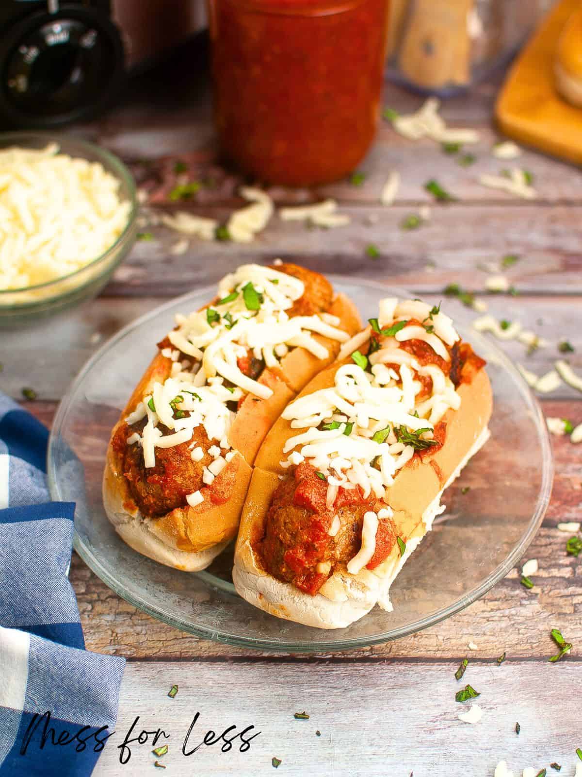 Two meatball subs on a glass plate, topped with melted cheese and garnished with chopped herbs. The wooden table is scattered with extra cheese and herbs. A bowl of shredded cheese, a jar of sauce, and a blue napkin are in the background.