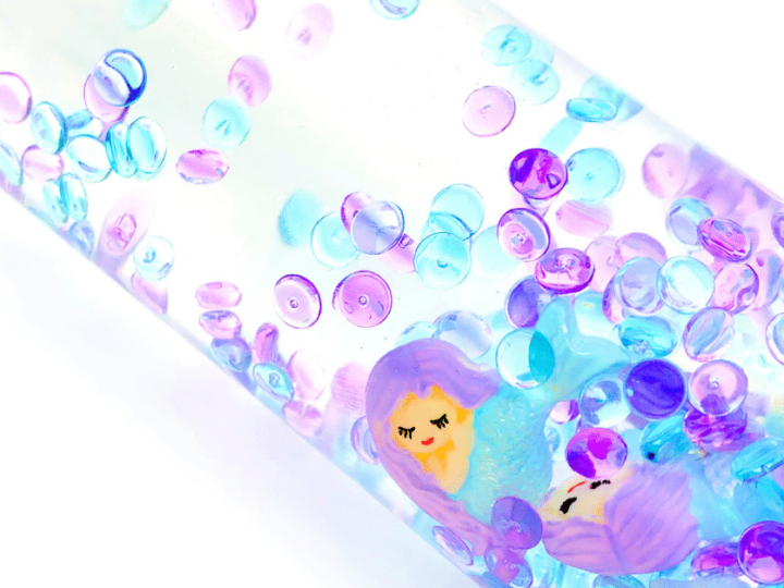 A sensory bottle filled with clear liquid contains colorful beads in shades of blue, purple, and pink. Two small mermaid figurines with purple hair float inside. The background is white. The image has Fun-A-Day written at the bottom left corner.