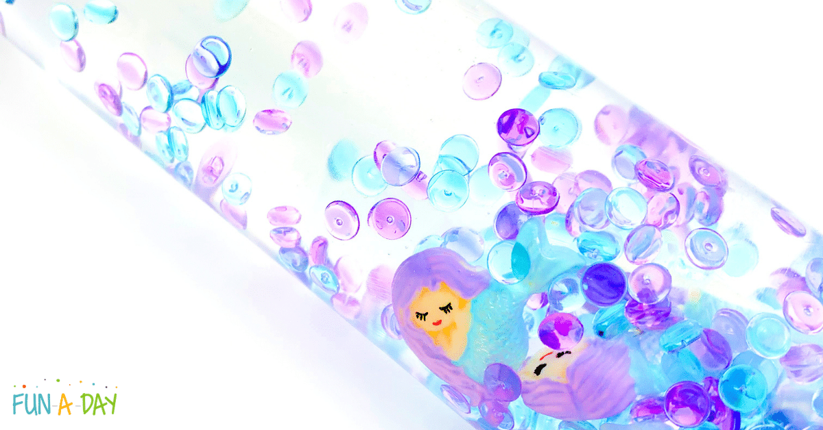 A sensory bottle filled with clear liquid contains colorful beads in shades of blue, purple, and pink. Two small mermaid figurines with purple hair float inside. The background is white. The image has Fun-A-Day written at the bottom left corner.