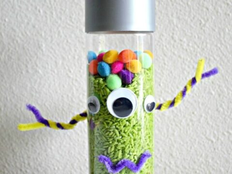 A transparent bottle with googly eyes, colorful pom-poms, and pipe cleaners creates a monster-themed discovery bottle. The cap is silver, and the bottle is filled with green rice. The text at the bottom reads MONSTER discovery bottle.
