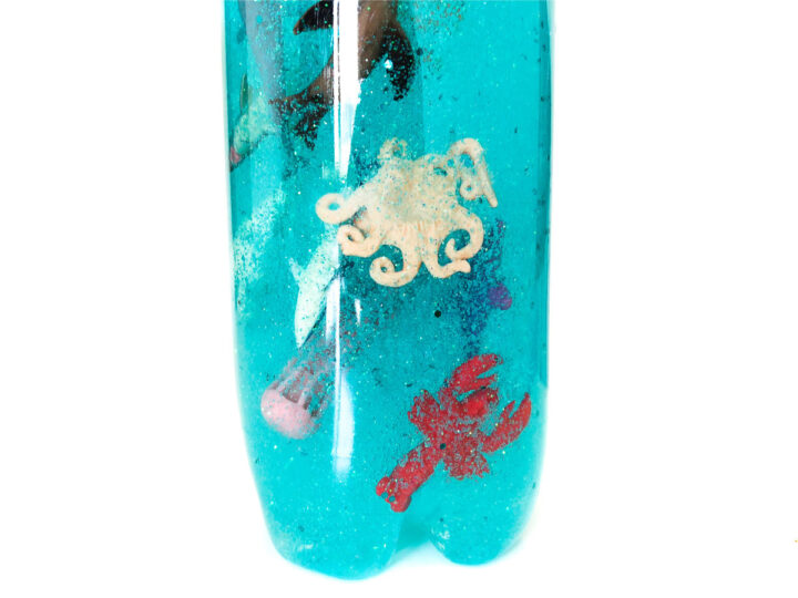 A close-up of a plastic bottle filled with blue liquid. Inside, there are floating plastic sea creatures, including an octopus and a red lobster. The bottles bottom indents are visible.